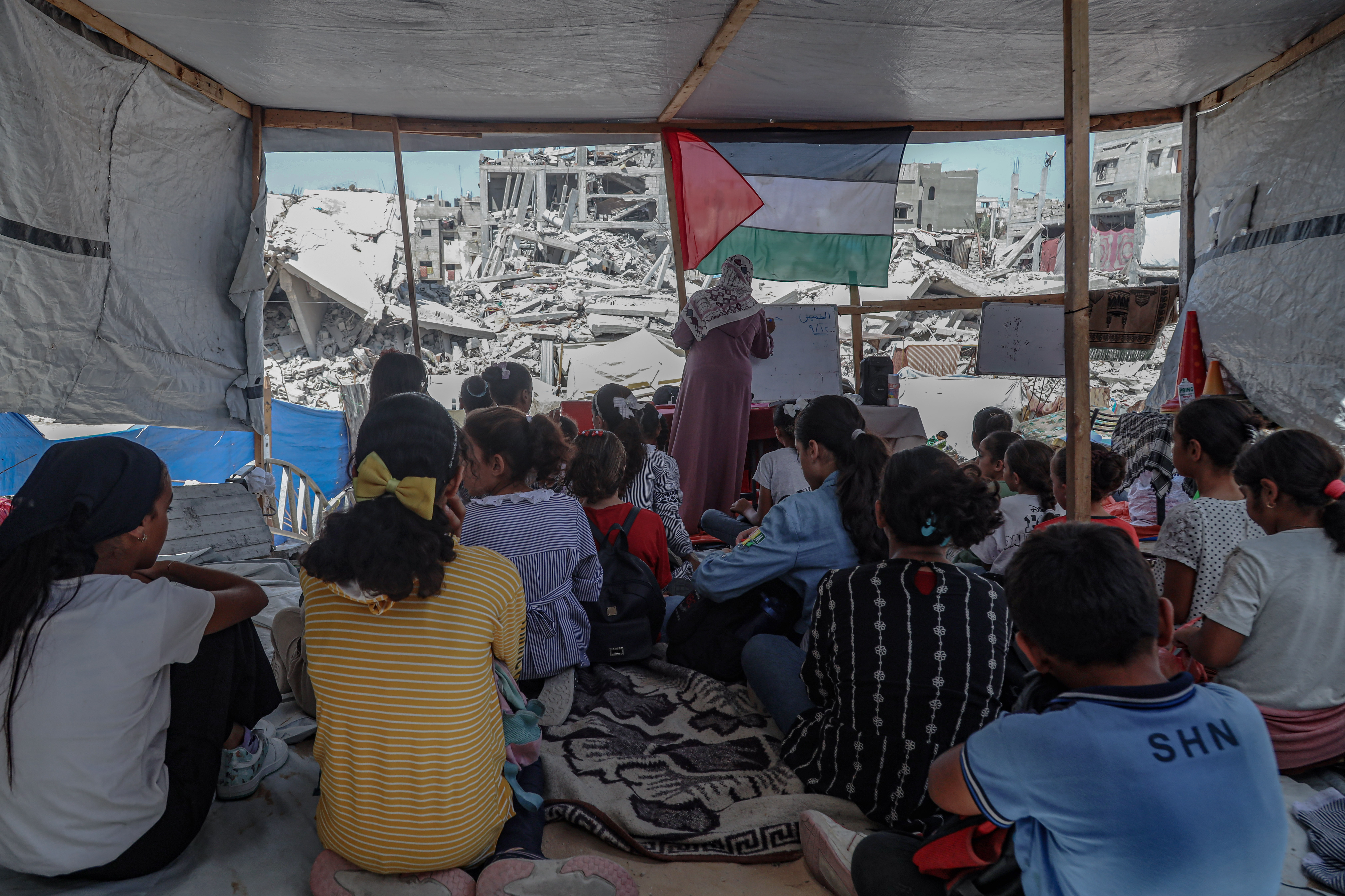 Eine Gruppe von Kindern sitzt auf dem Boden eines zerstörten Hauses. Vorne steht eine Lehrerin, hinter ihr eine palästinensische Flagge und ein Blick auf ein zerstörtes Gebäude.