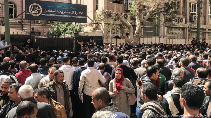 Hundreds of people stand close together in front of a large stone building, some of them wearing masks