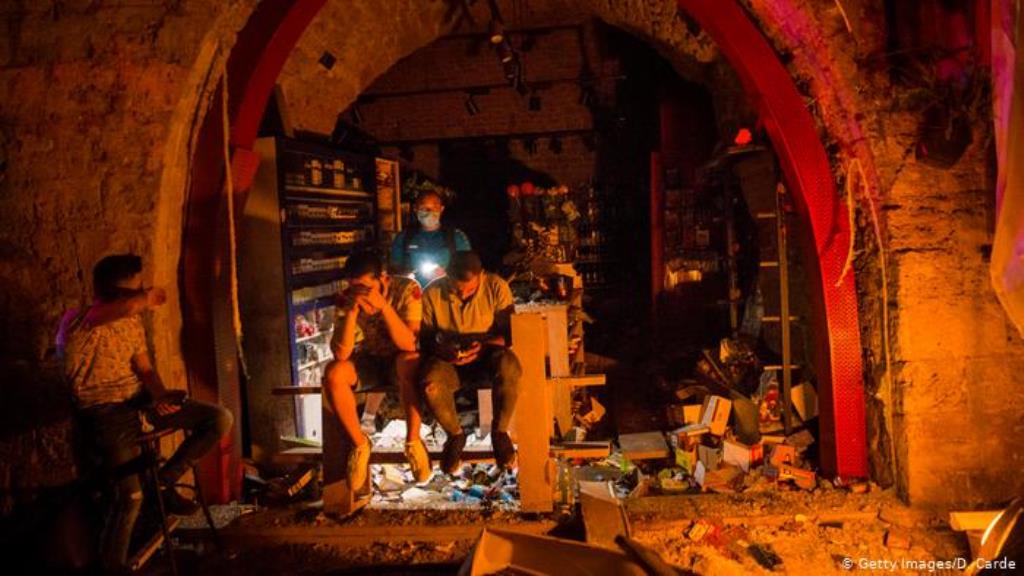 Ein paar Männer sitzen im Halbdunkel vor einem zerstörten Laden, einer checkt sein Telefon (Getty Images/D. Carde)