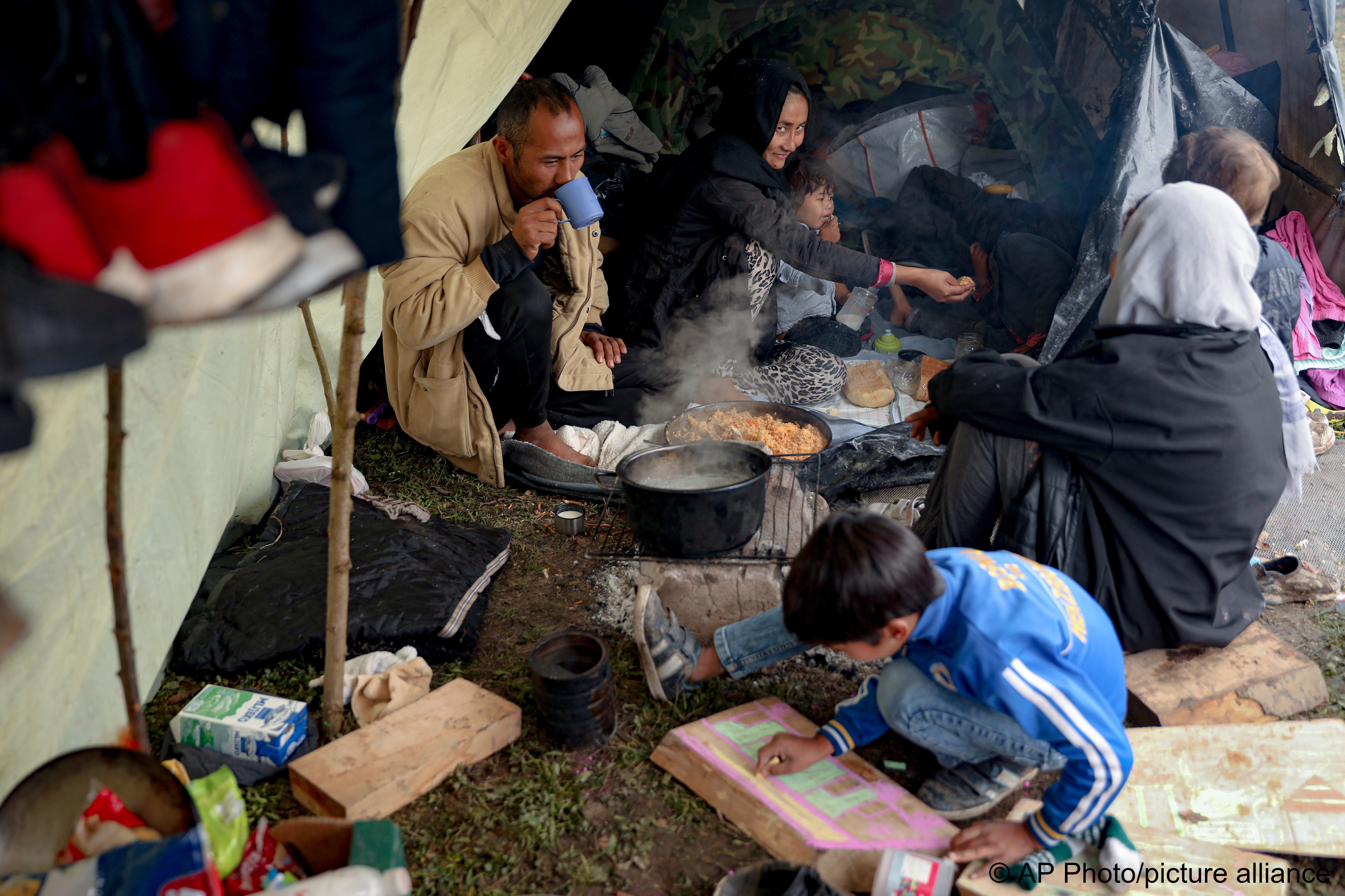 Ein Migrantenkind malt auf einem Stück Holz, während die Menschen essen in einem behelfsmäßigen Lager für Migranten, hauptsächlich aus Afghanistan, in Velika Kladusa, Bosnien, am 12. Oktober 2021; Foto: AP Photo