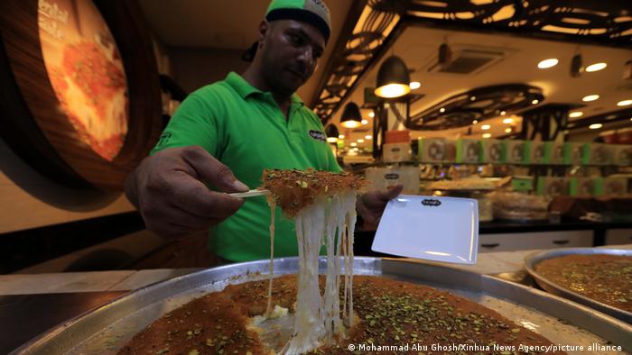 Man dripping stick over a large platter of a dark gooey dessert with crushed pistachios