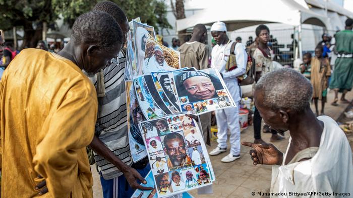 Three men in the foreground look at posters of religious personalities, while others in the back look into their phones