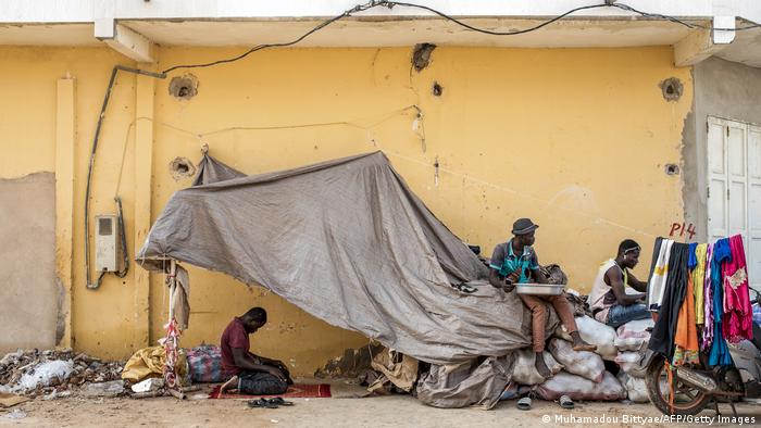 A man prays beneath an improvised roof made out of tarp, as two other men sit on sacks next to a motorcycle behind a laundry line with bright clothes hanging from it