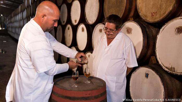 Zwei Rum-Meister stehen in weißen Kitteln vor Fässern, einer der beiden schenkt Rum in ein Glas ein