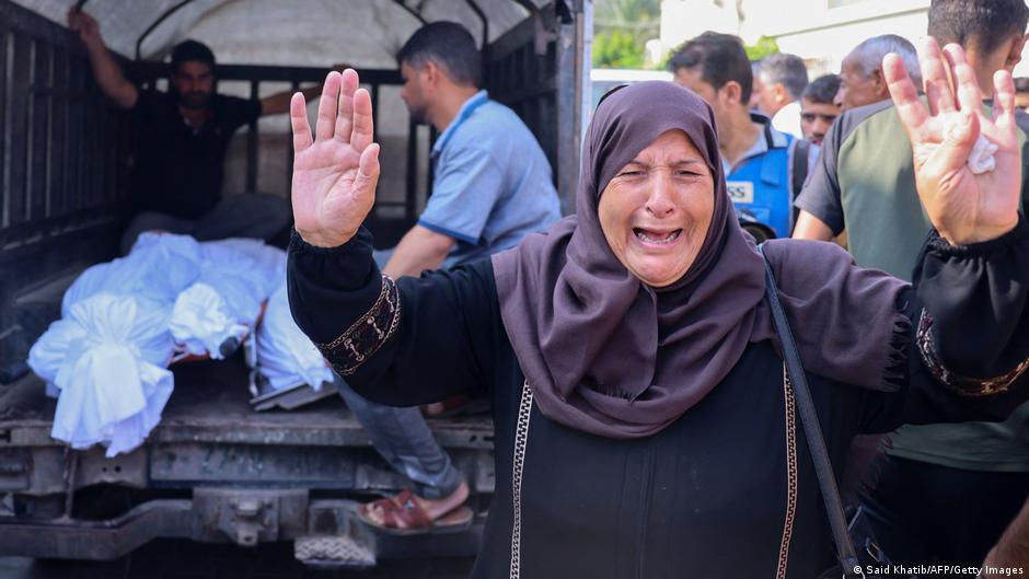 A woman wails as she raises her hands, as men load blanketed bodies onto a truck in the background