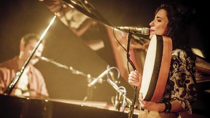 Djazia Satour (right) sings into a microphone as she plays the bendir; in the background, sitting at a grand piano, is pianist Pierre-Luc Jamain