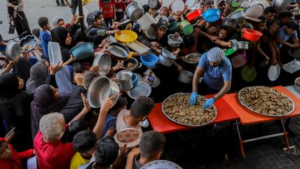 Eine große Gruppe von Menschen mit leeren Tellern, Schüsseln und anderen Gefäßen drängt sich um einen Helfer, der Essen aus einer großen Pfanne ausgibt.