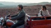 A man sits astride a motorbike while a woman sits in the trailer behind, against a background of hilly dry terrain 