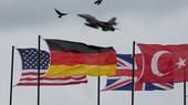 American, German, English and Turkish flags are waving in the wind. A NATO fighter jet flies above them.