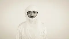 A man (Nigerien Musician Oumara Moctar/Bombino) dressed entirely in a white traditional Tuareg outfit and headdress against a white backdrop